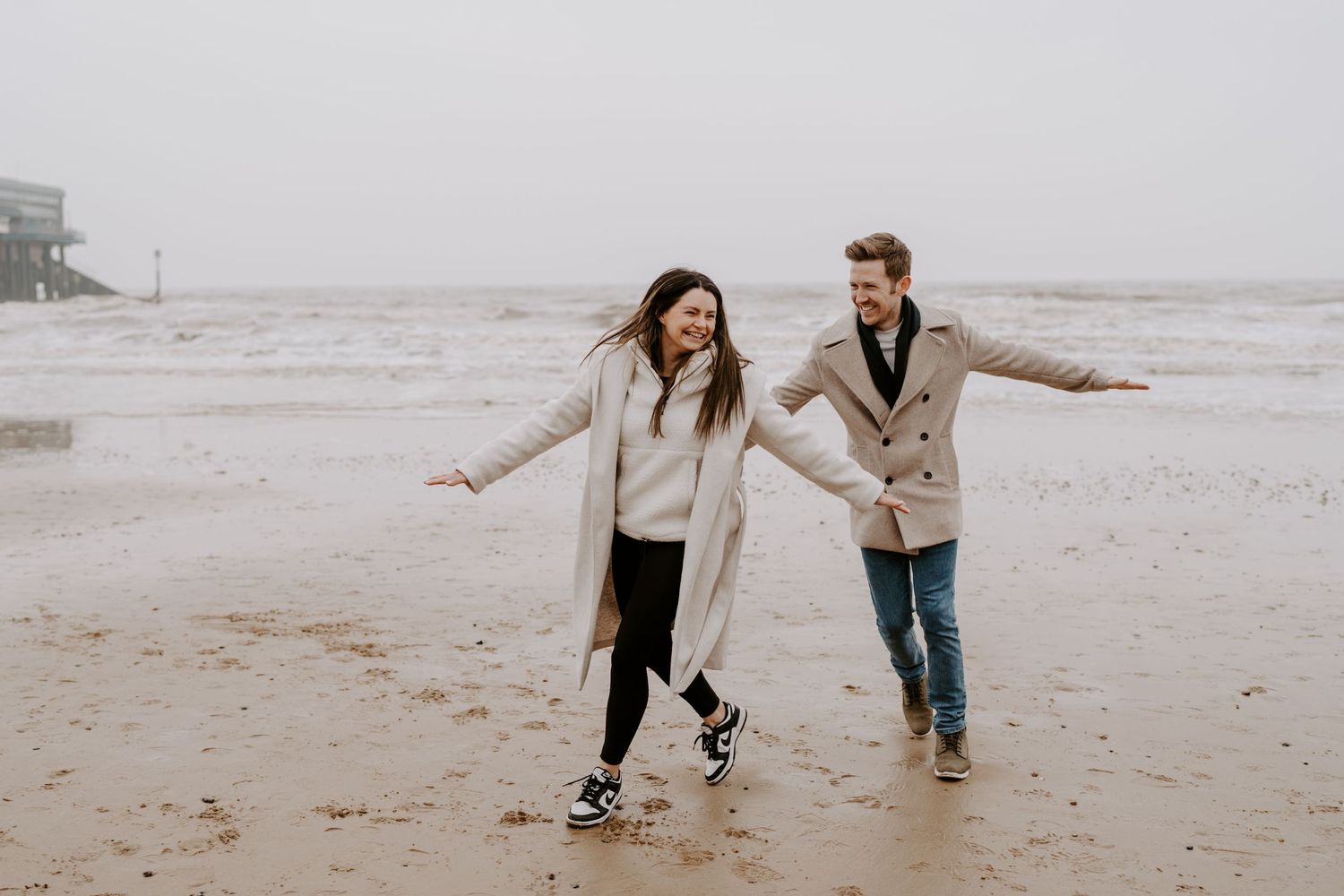 A couple are running along the beach with their arms outstretched and laughing