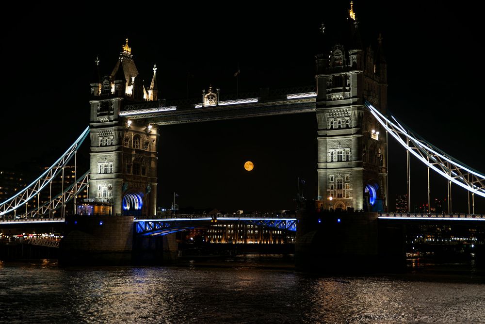 Night view of London's Tower Bridge illuminated in blue against dark sky with reflections on the River Thames.