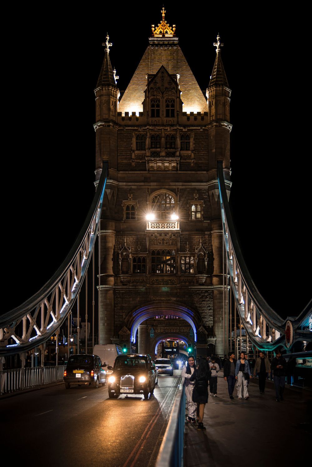 Night view of Tower Bridge in London illuminated with warm lights as vehicles and pedestrians cross the historic structure.