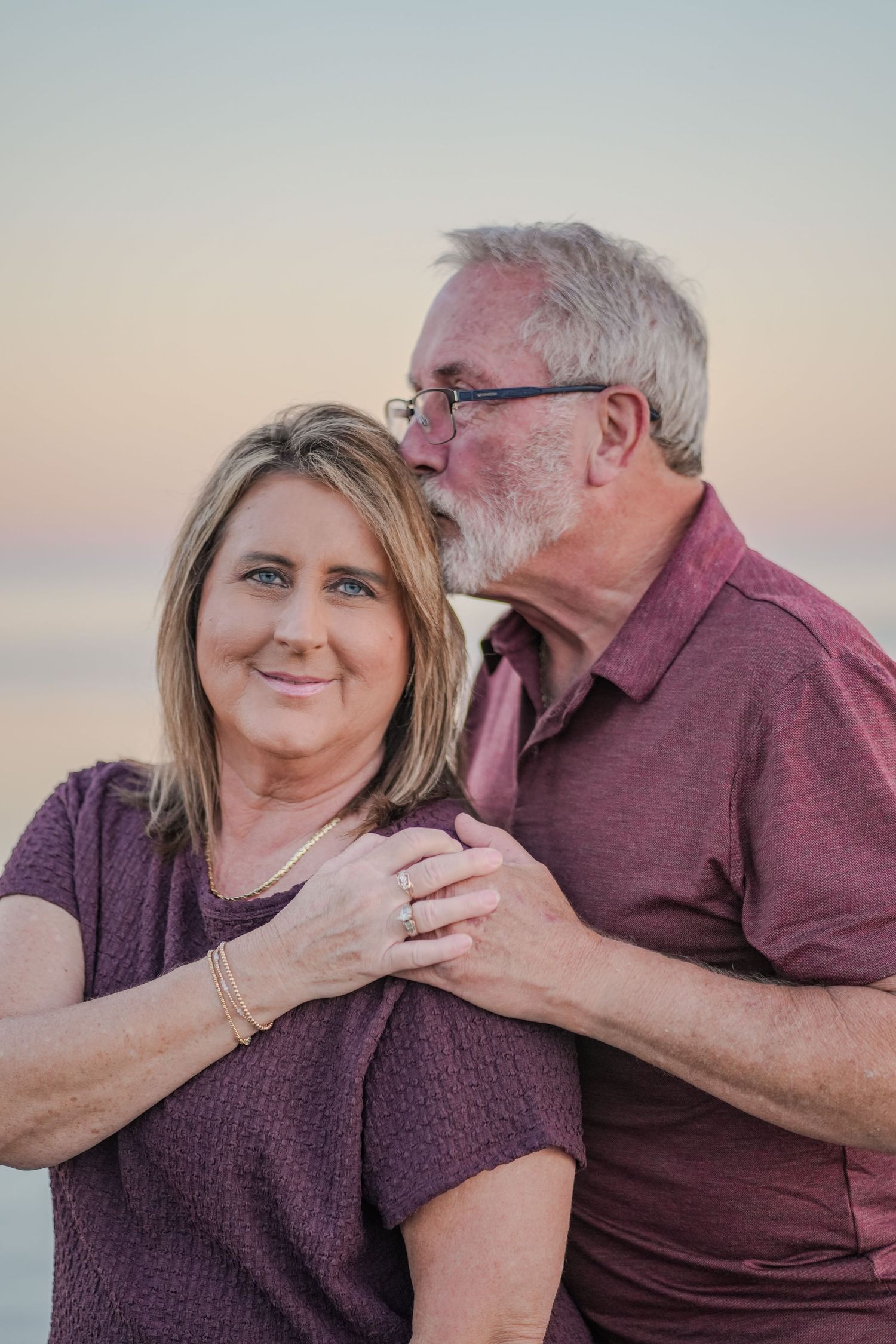 Older man kissing woman on cheek at sunset beach, both wearing purple.