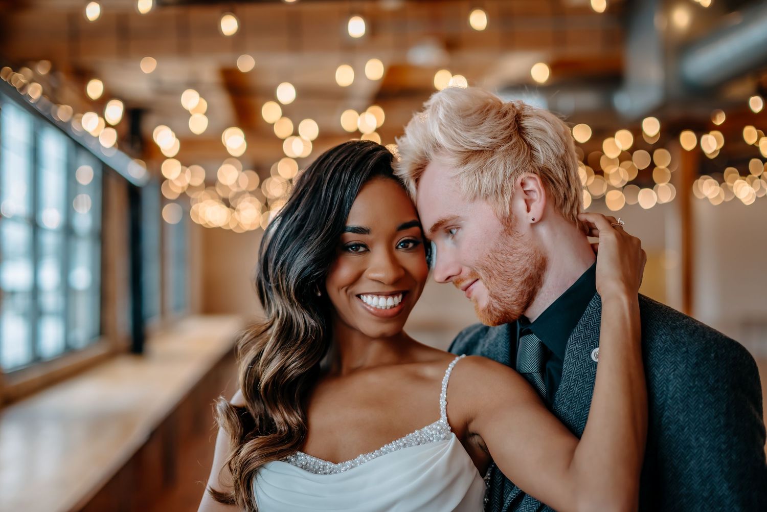 Couple share an intimate moment laughing together under warm string lights in an indoor venue.