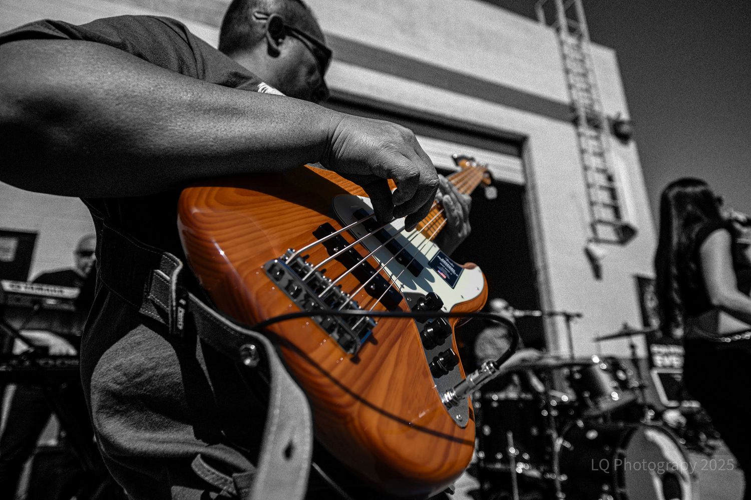 Selective color image of hands playing an orange bass guitar during outdoor performance.