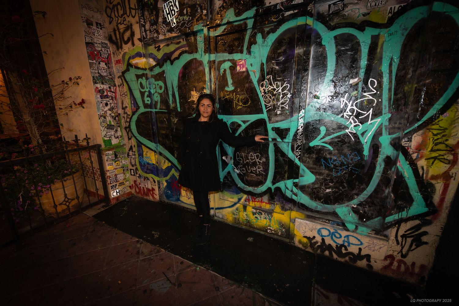 Figure in black poses against colorful graffiti wall with turquoise lettering in dark urban setting.