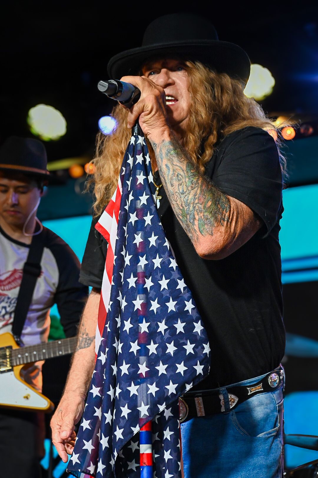 A performer wearing an American flag vest sings into a microphone on stage during a concert.