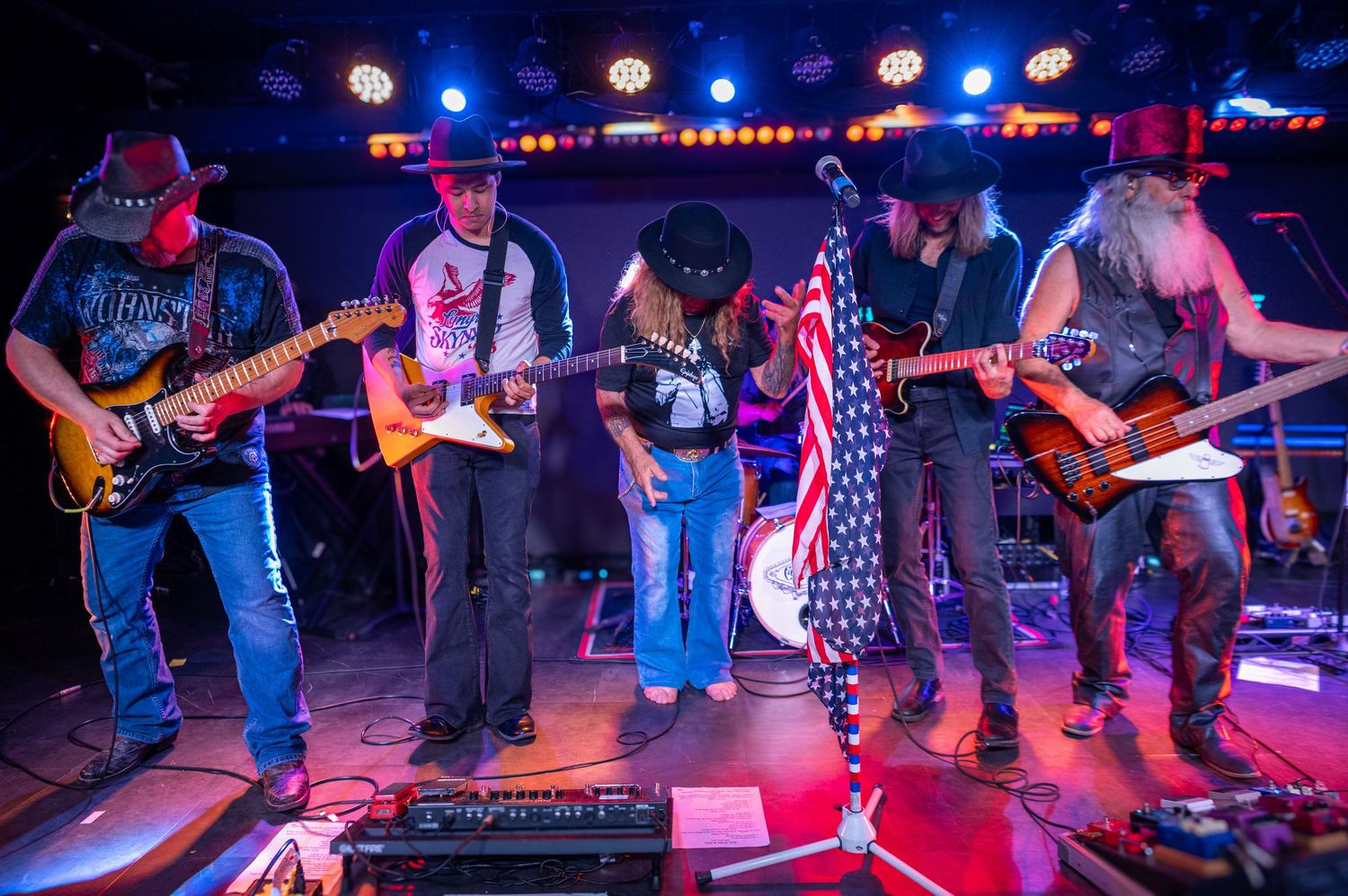 Musicians playing guitars together on stage with American flag and blue and red lighting.