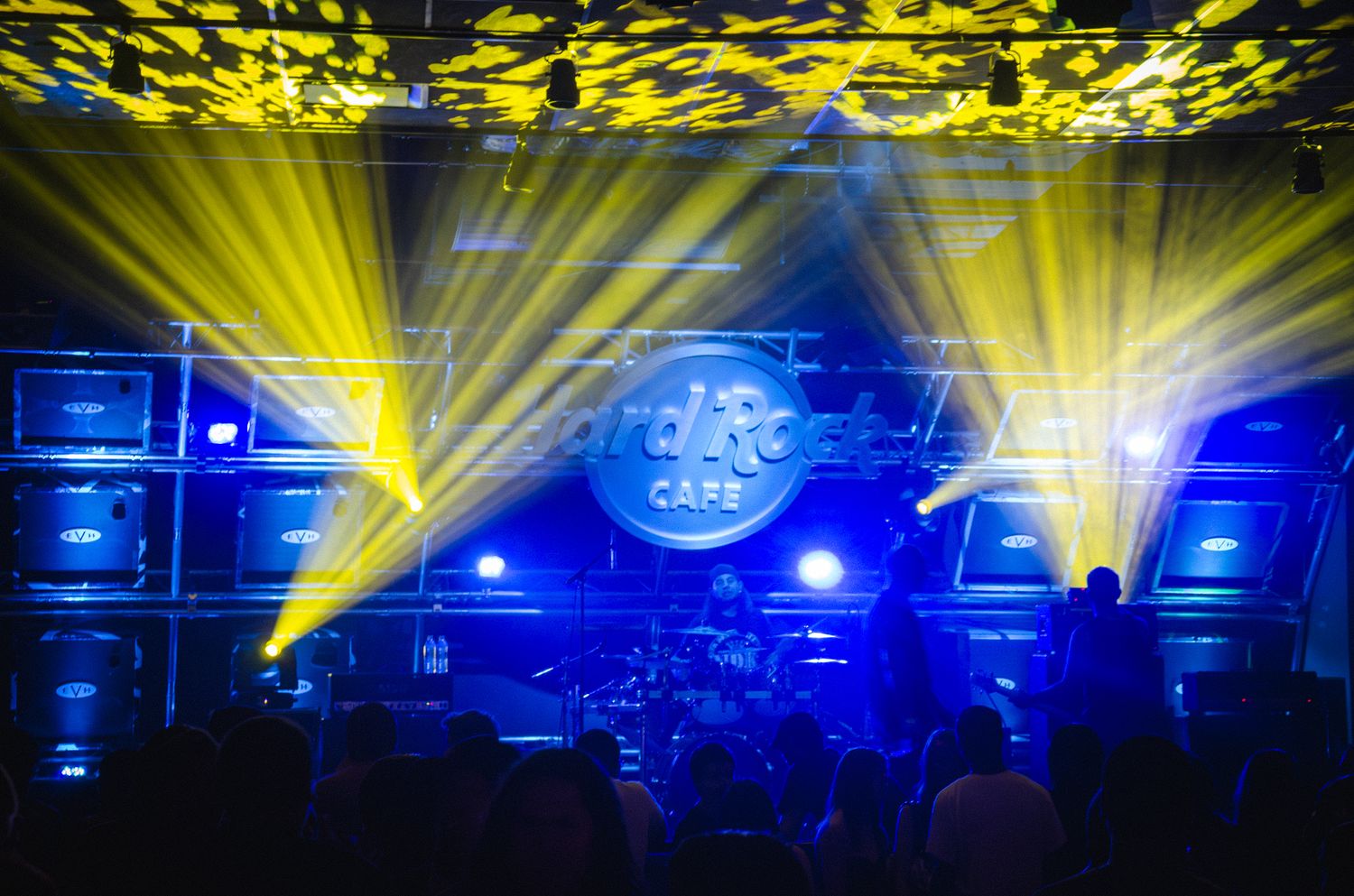 Concert stage lit with dramatic yellow spotlights and blue backlighting against wall of amplifiers.