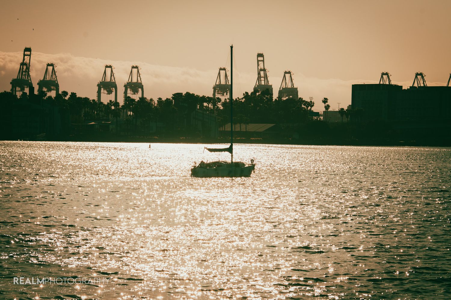 Silhouette of sailboat against sunset with industrial cranes on harbor skyline.