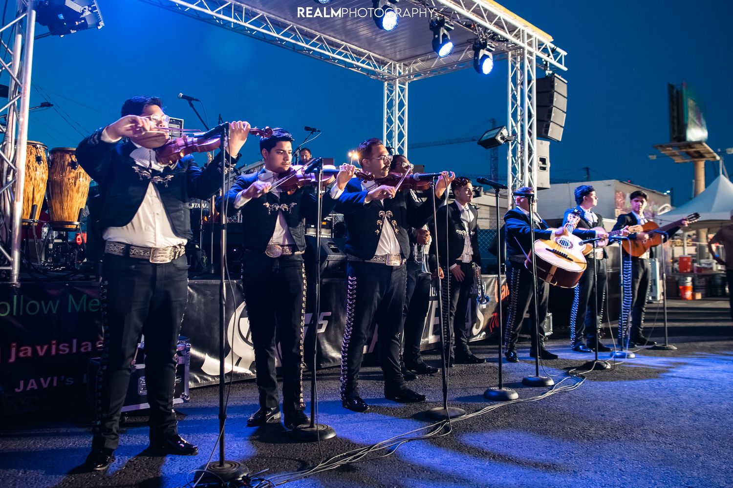Large mariachi band performing on outdoor stage with lighting equipment.
