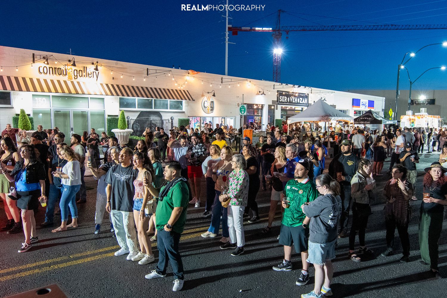 Large crowd of people gathered outside storefronts at night with street lights illuminating the scene.
