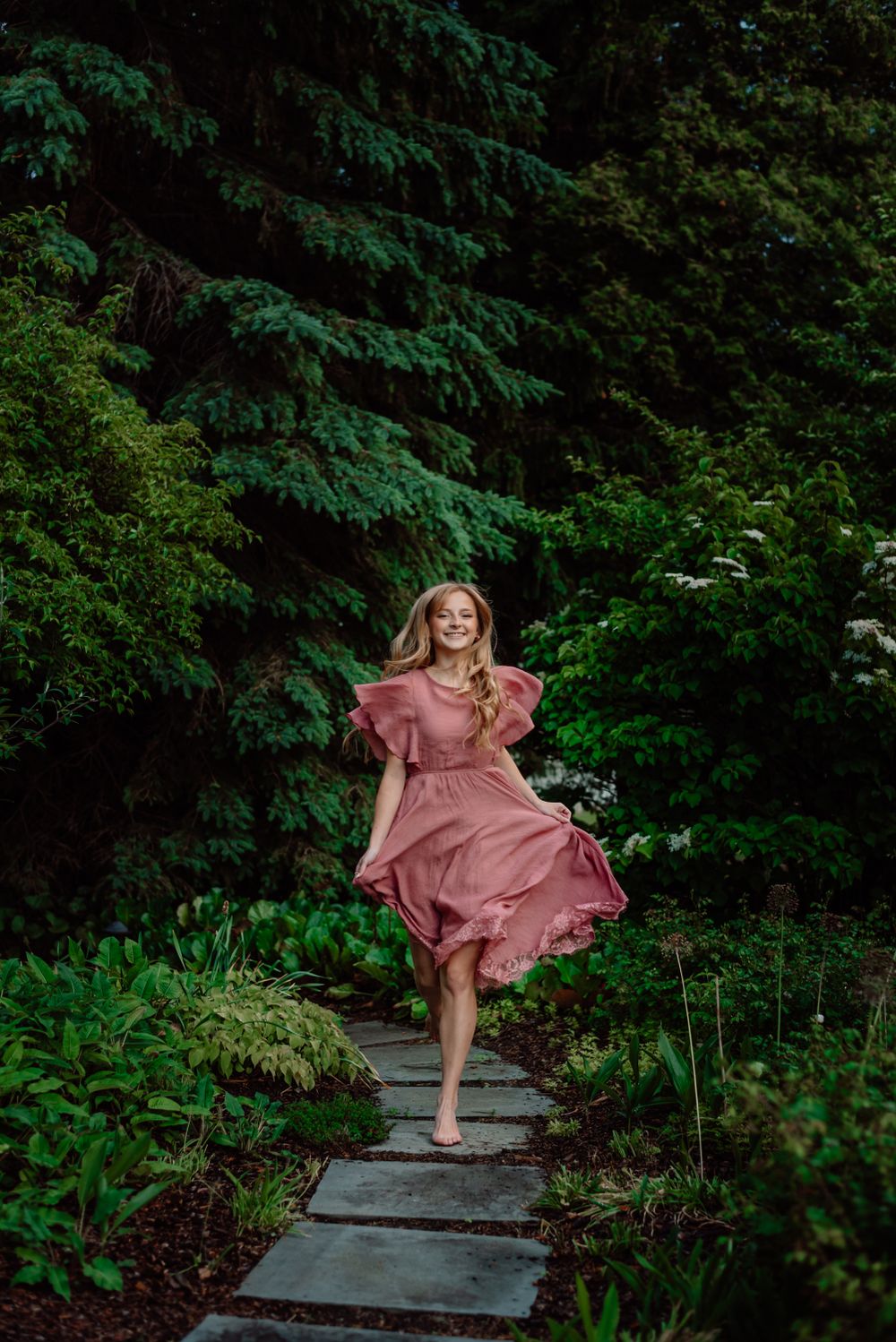 A flowing pink dress moves gracefully along stone steps in a lush green forest setting.