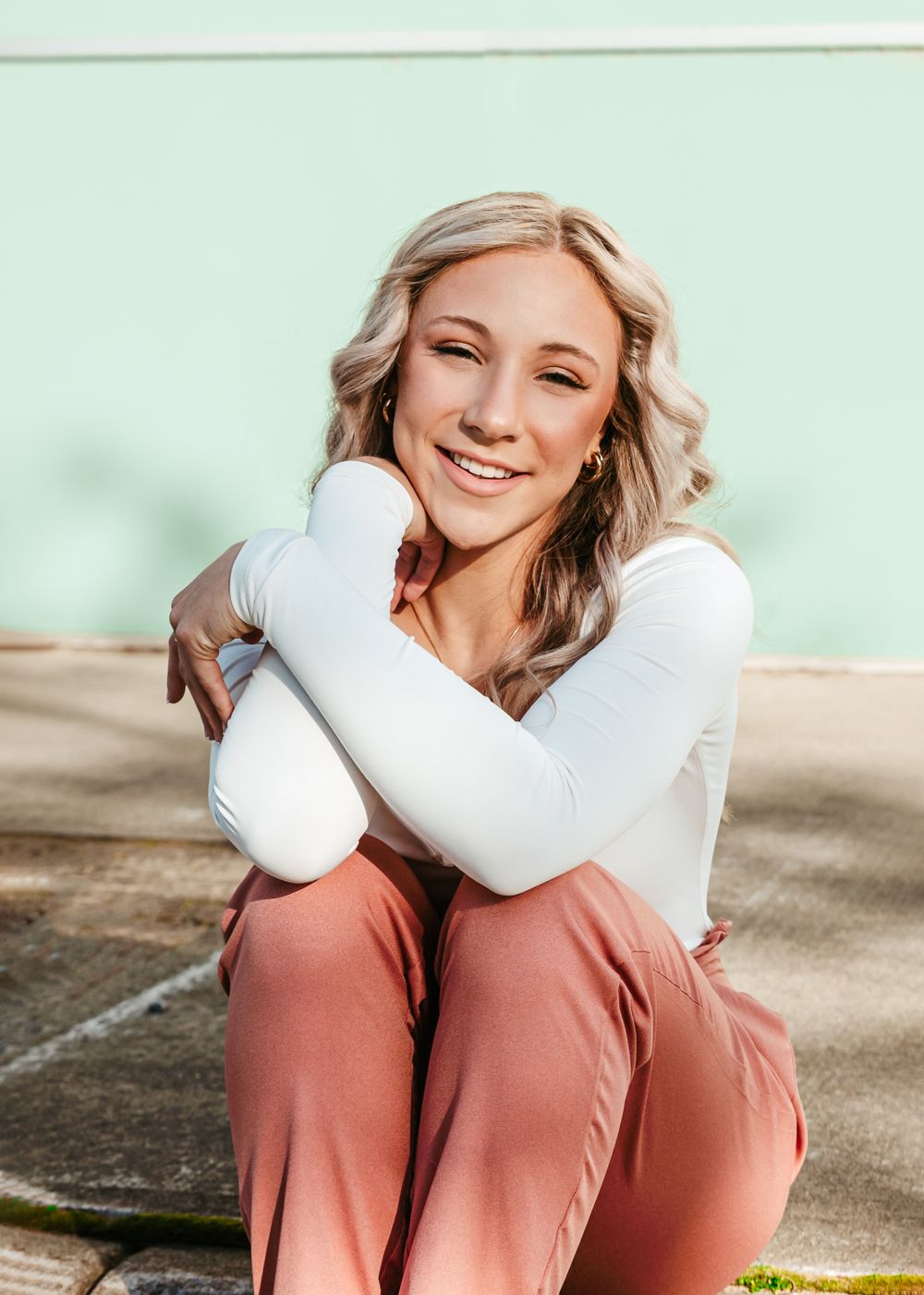 Portrait of someone in casual attire with a white long-sleeve top and rust-colored pants sitting outdoors against a mint wall.