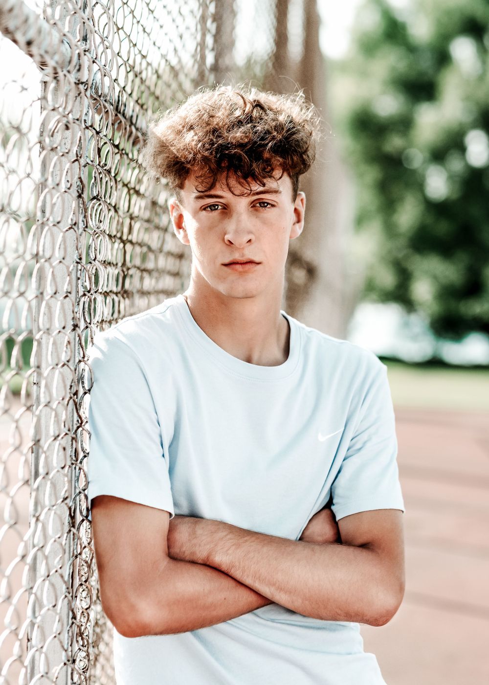 A person with curly hair wearing a white t-shirt leans against a chain link fence outdoors.