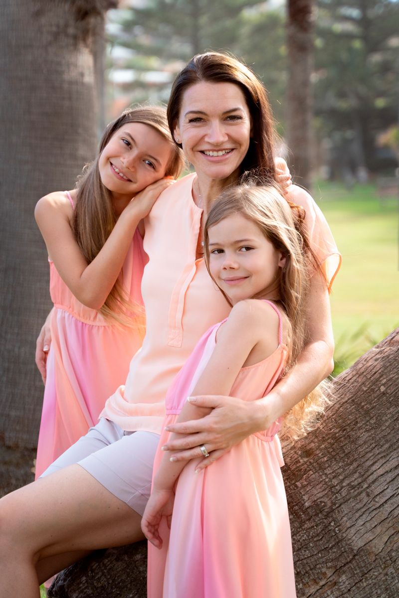 Mom sitting on a palm tree in the park at freshwater beach hugging her daughters during a family photoshoot in the northern beaches Syndye