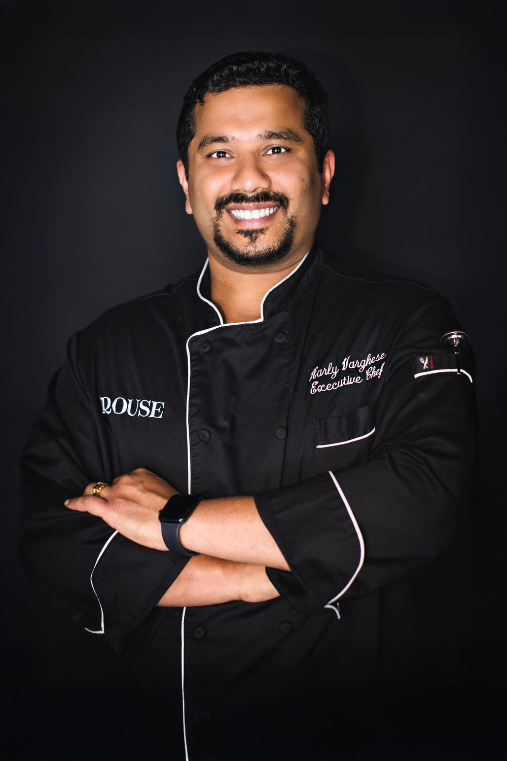 Professional chef poses in black uniform with white piping against dark background in dramatic lighting series.