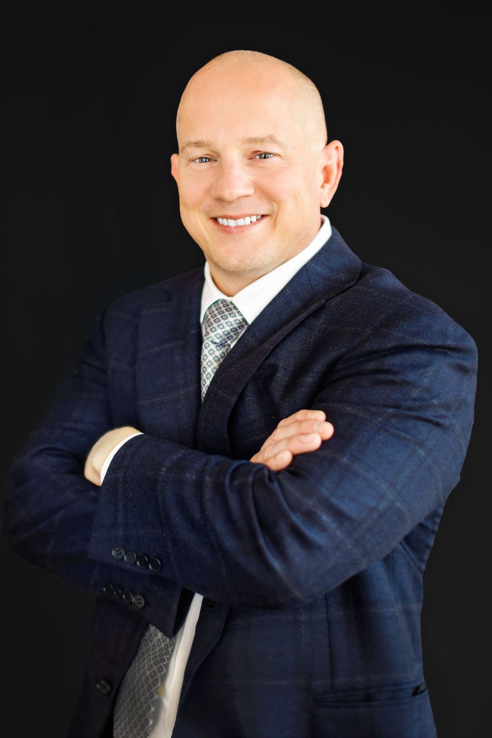 Professional headshot of a person in a navy suit and gray tie with arms crossed against dark background.