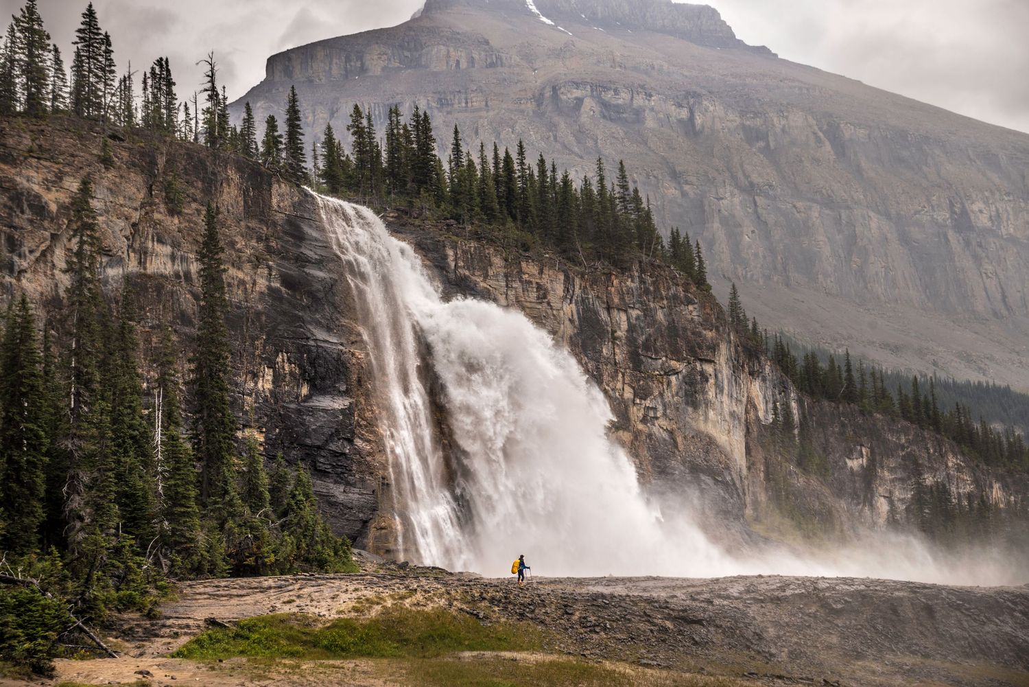Powerful waterfall cascades down rocky cliff face surrounded by evergreen forest and mountain backdrop.