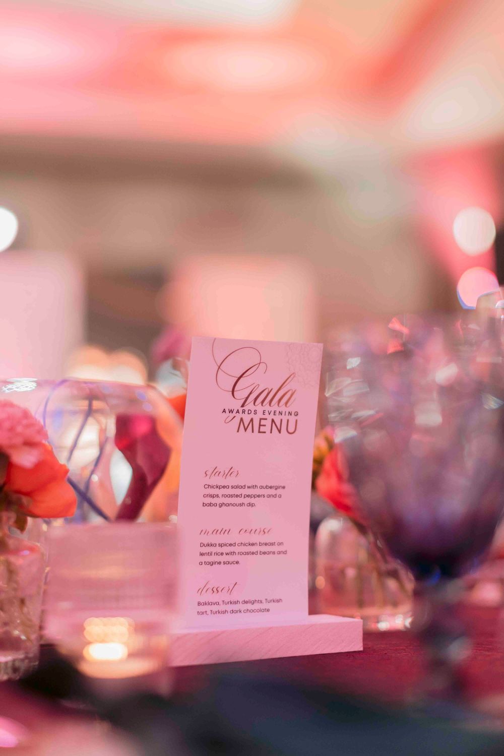 Pink Gala Awards Evening menu card displayed in a holder surrounded by colorful flowers and candlelight on a table.