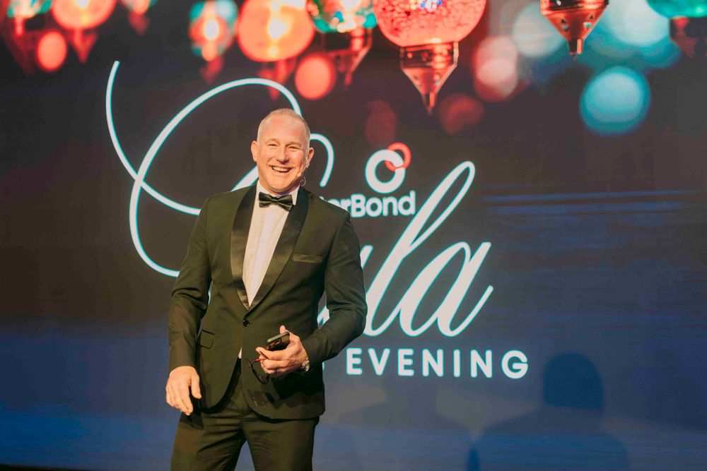 Confident man in tuxedo hosting BetterBond Gala Awards Evening on illuminated stage.