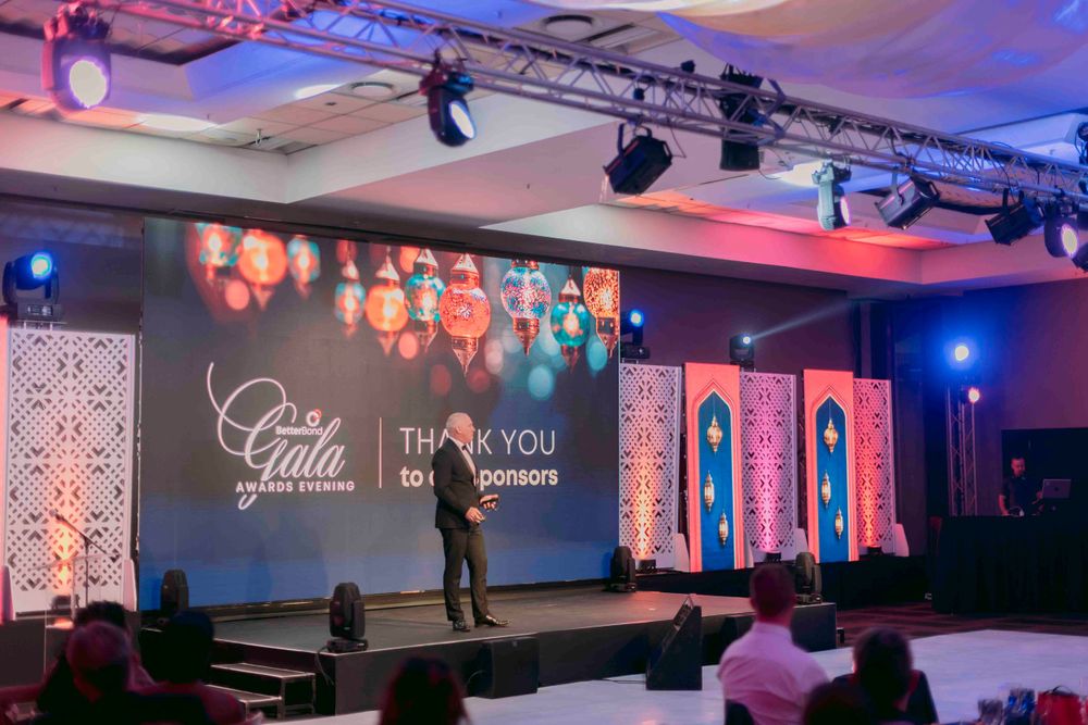 Speaker on stage at BetterBond Gala Awards Evening with thank you sponsors message on large screen.