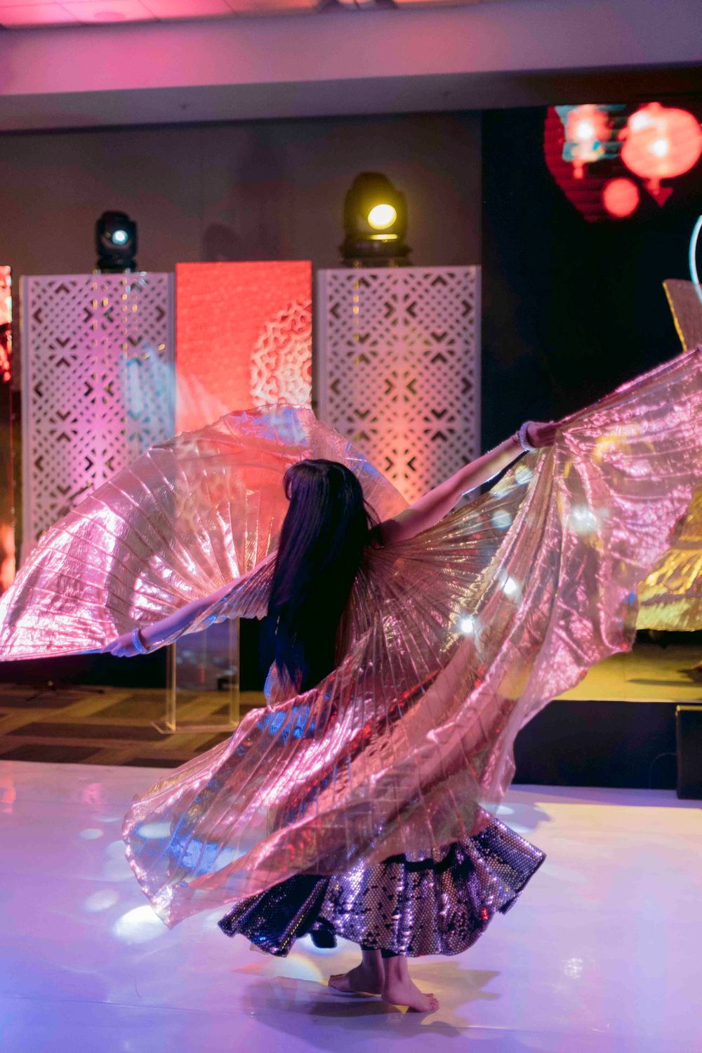 Belly dancer performing with illuminated pink iridescent isis wings on a stage with colorful event lighting.