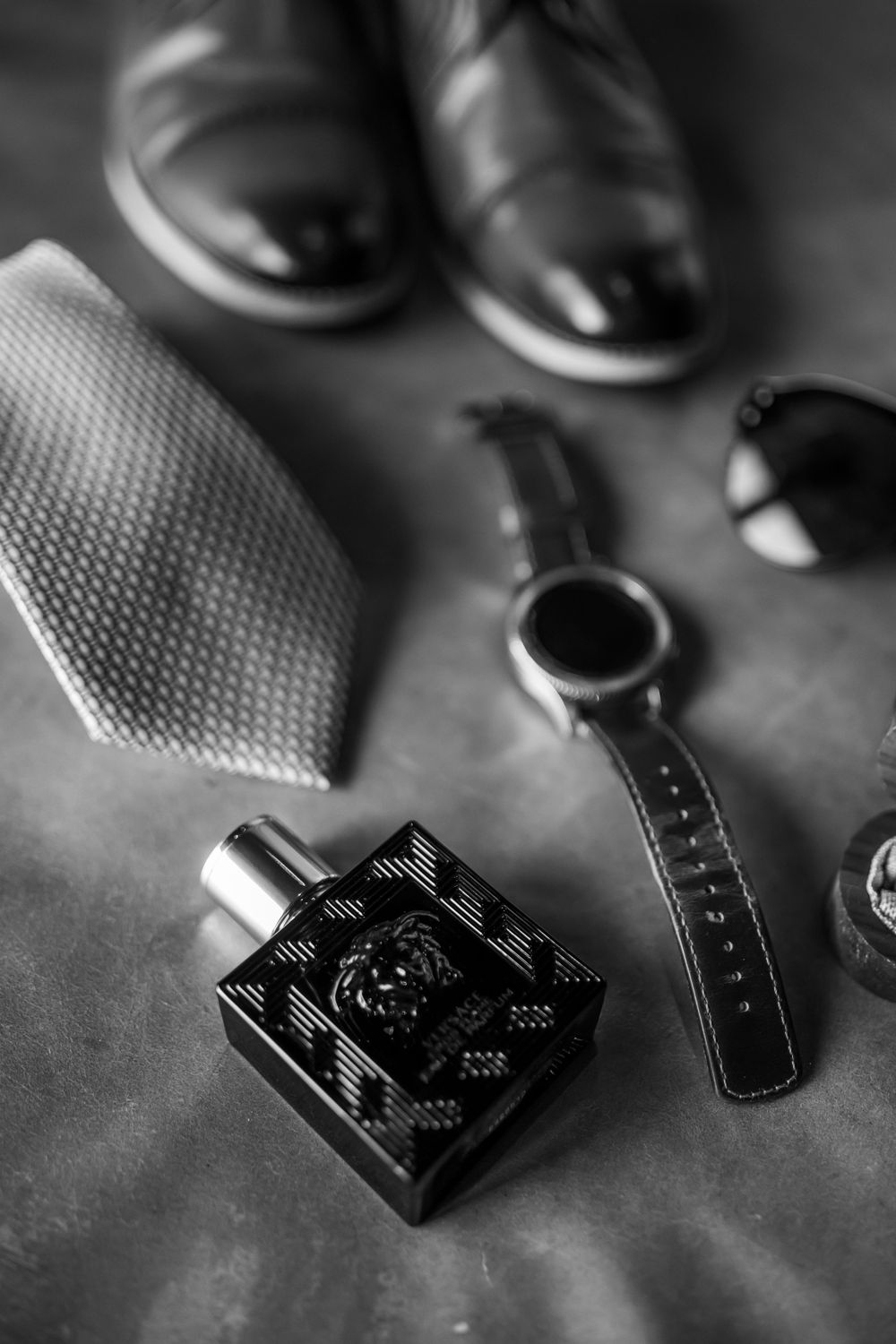 Wedding accessories including dress shoes, tie, watch and cologne bottle arranged in black and white.