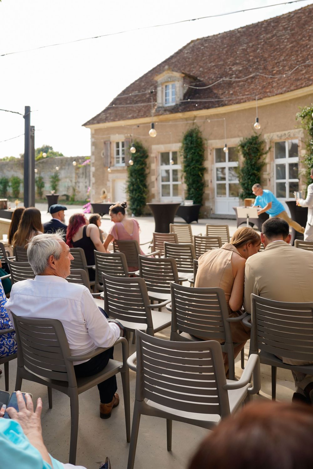 Audience members sitting in rows of modern chairs on a patio facing a stone building with climbing vines.