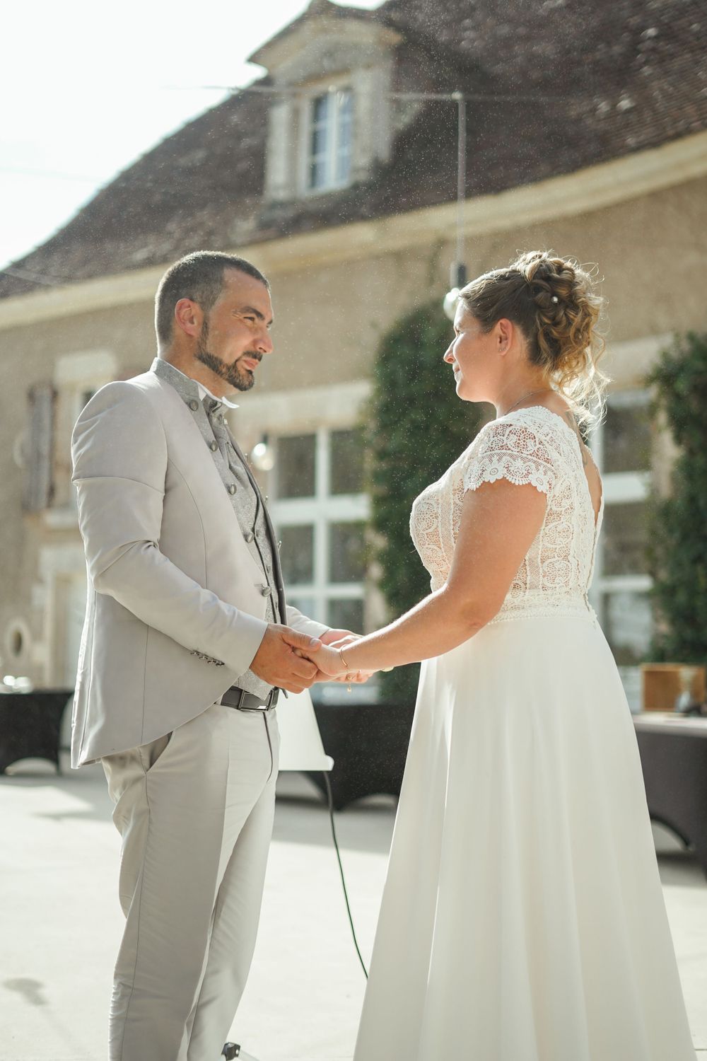 A wedding couple holds hands during their outdoor ceremony in front of a rustic stone building.