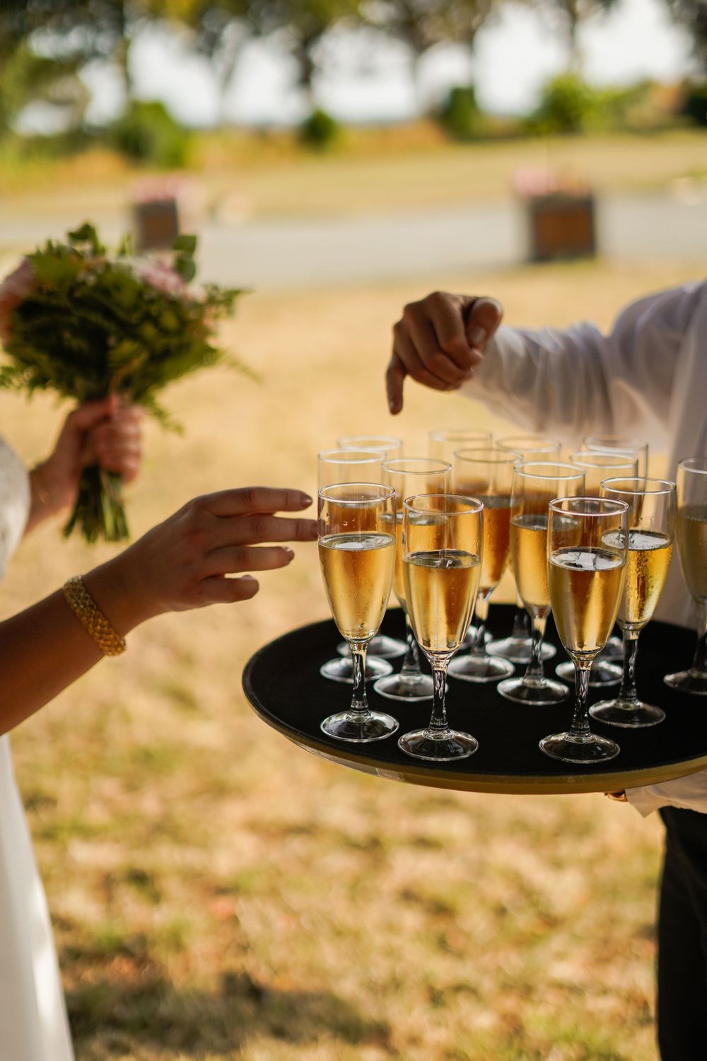 Server presents tray of champagne flutes while hand reaches for glass during outdoor reception.