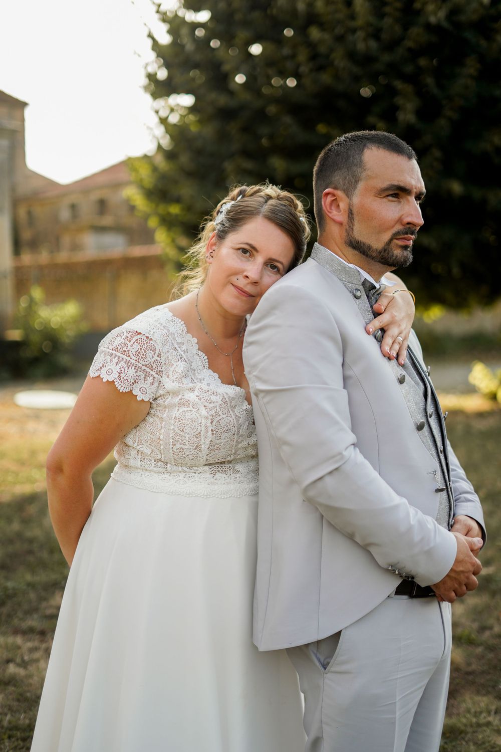 A tender wedding portrait of a couple in white and light gray attire against a sunlit garden backdrop.