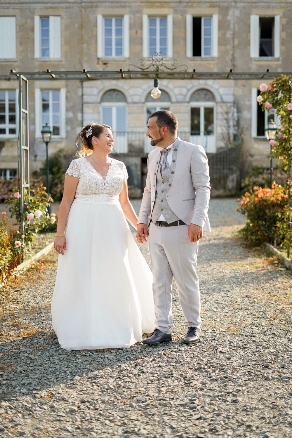 Newlyweds share a moment on gravel path in front of elegant stone mansion with climbing vines.