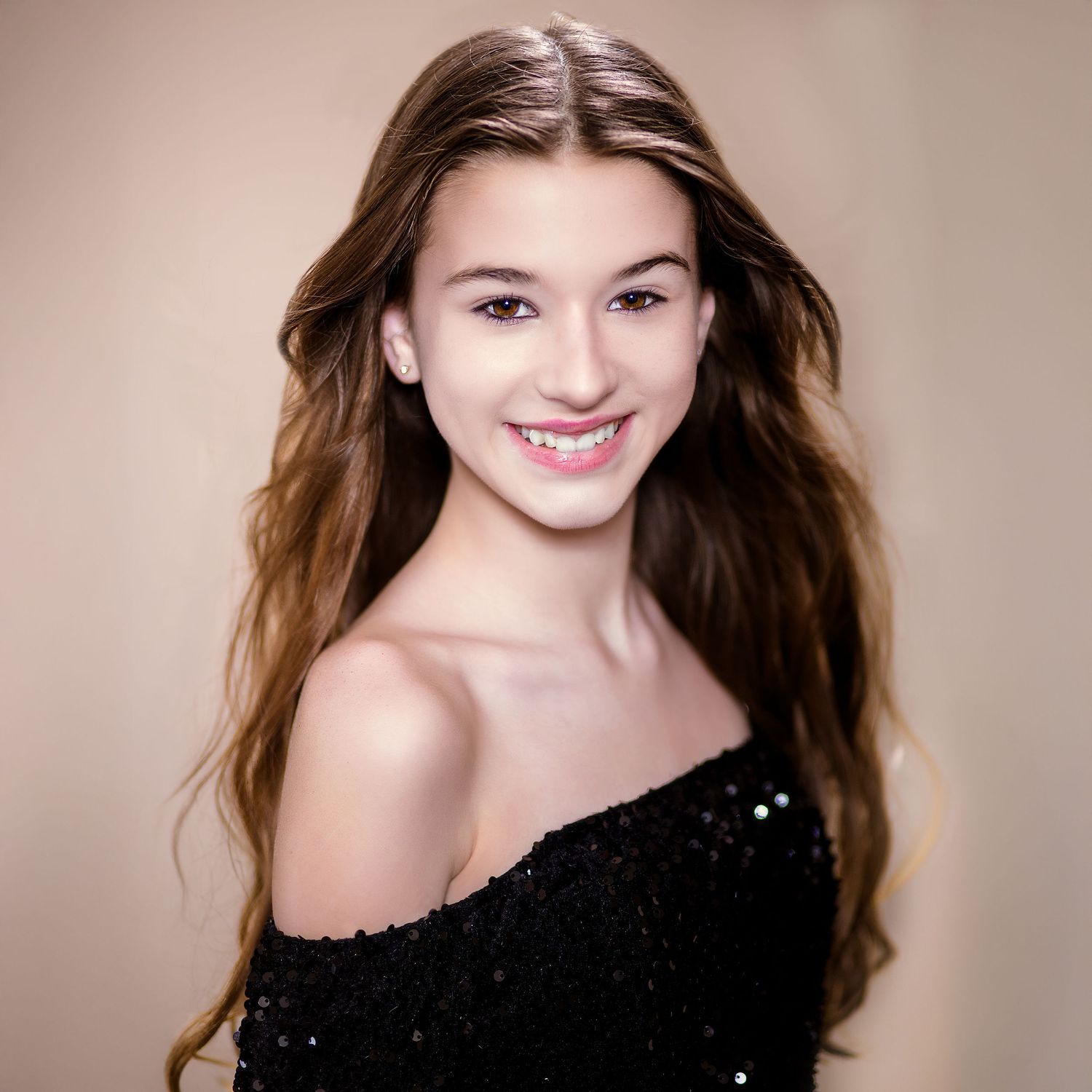 A smiling woman with long brown hair wearing an off-shoulder sparkly black top against a neutral background.
