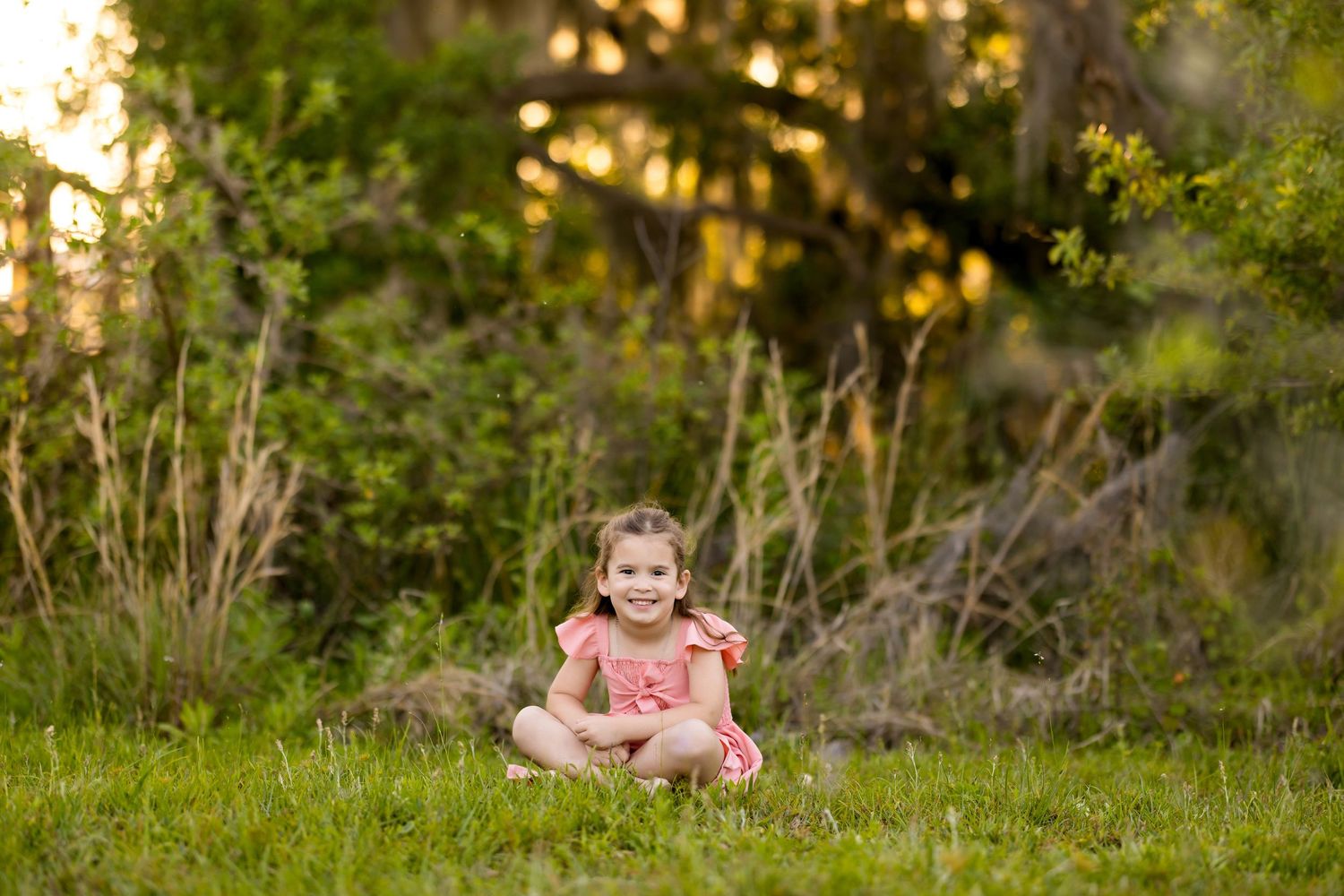 Small figure in pink dress sits cross-legged in sunlit meadow with trees in background. Photos by Tampa Photographer DigitalMyst Photography