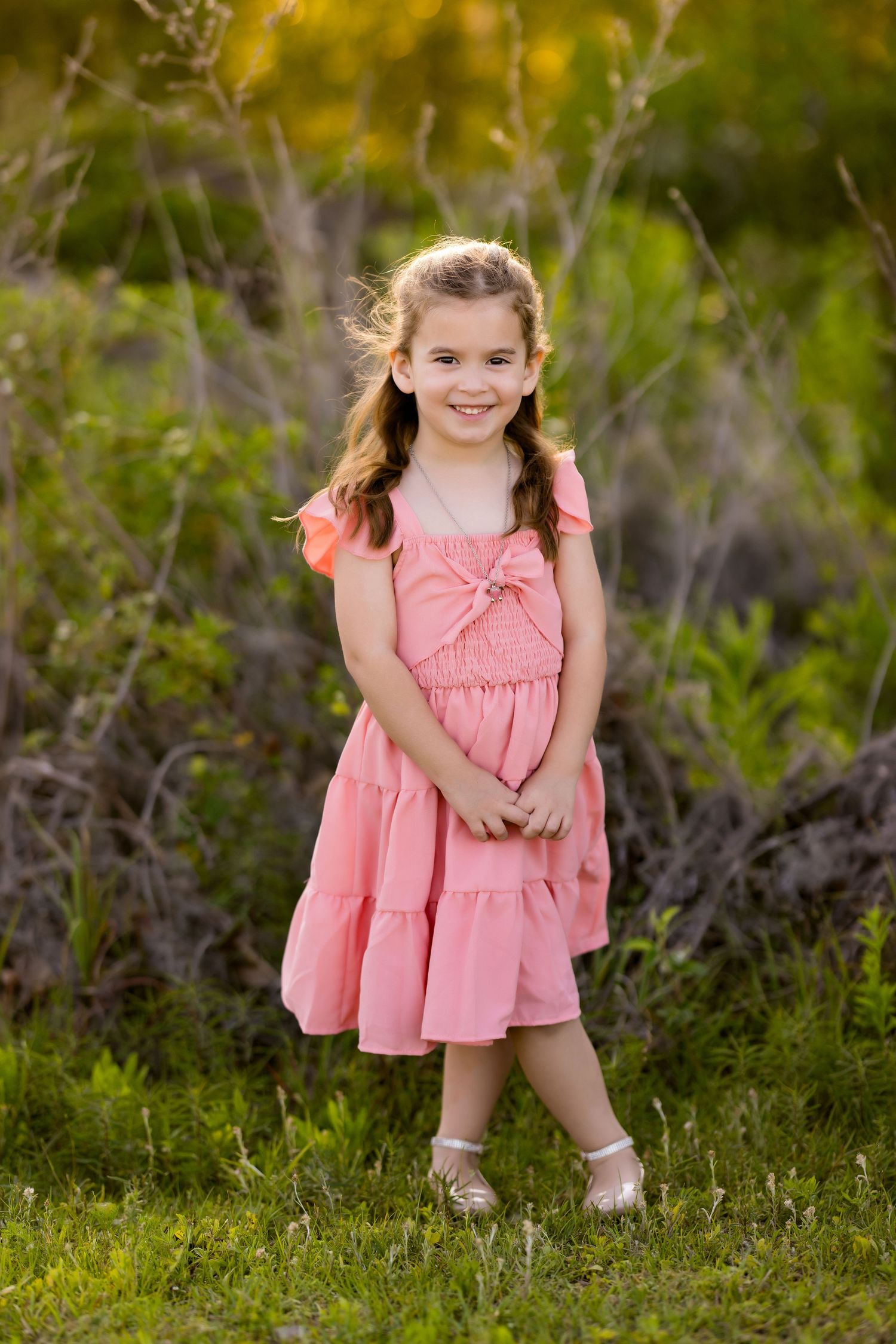 Pink tiered dress adorns a joyful pose against lush greenery in warm evening light.