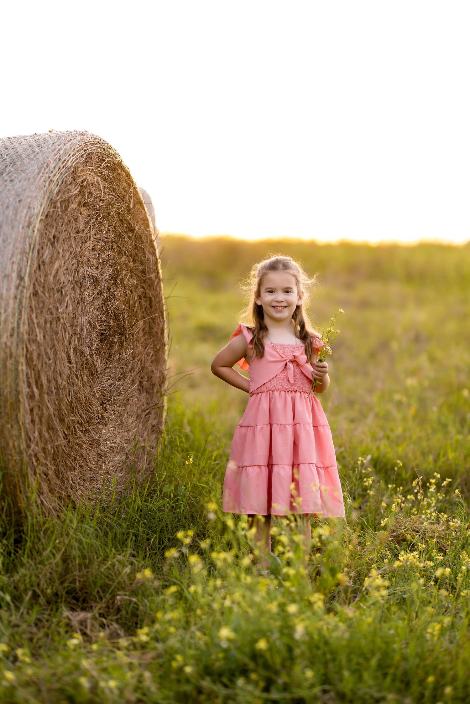 A young person in a pink ruffled dress stands next to a hay bale in a sunlit field of wildflowers.