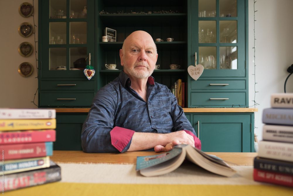 Someone sits at a wooden table with a stack of books while posed against teal-colored kitchen cabinets in the background.