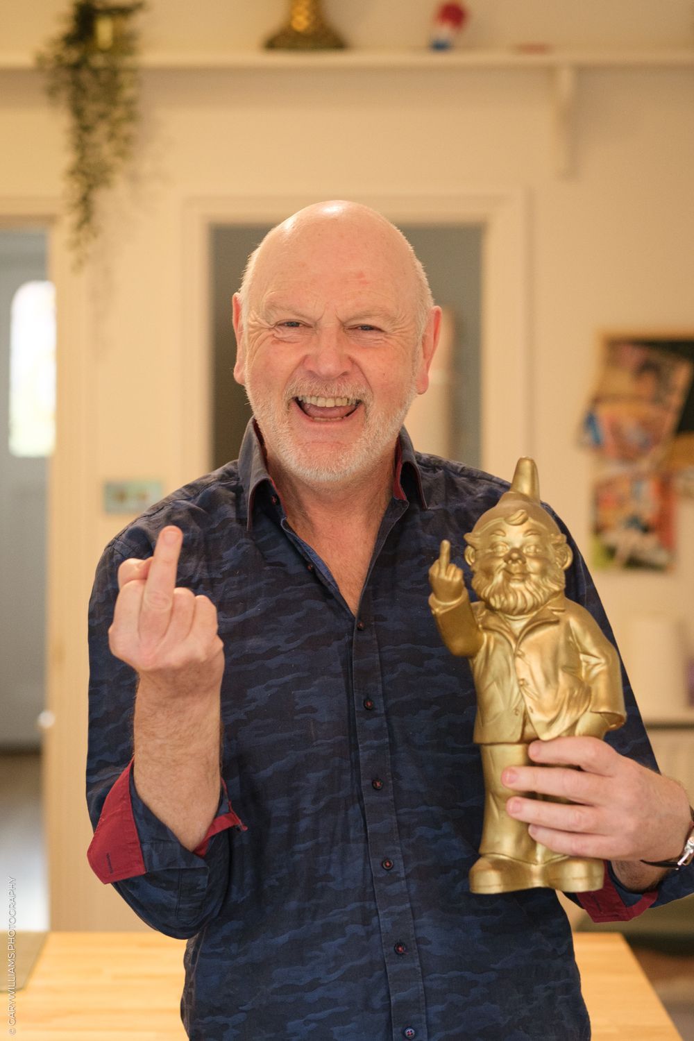A person displaying the middle finger while holding a golden garden gnome statue in a home interior.