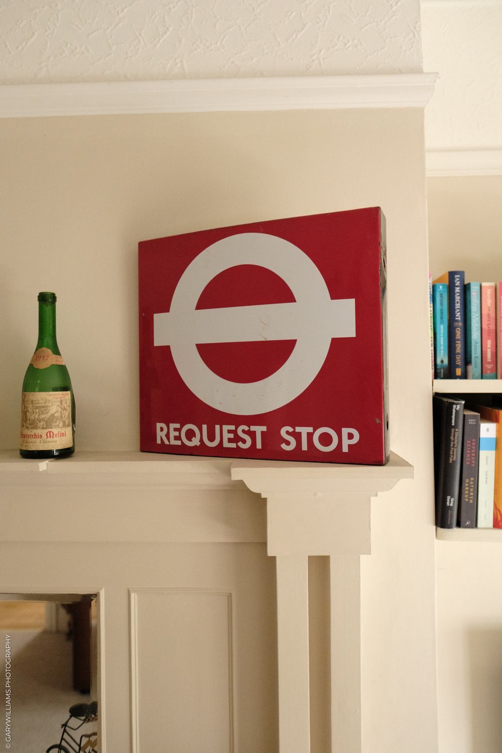 A vintage London Transport Request Stop sign hangs on a cream wall beside a green wine bottle and bookshelf.