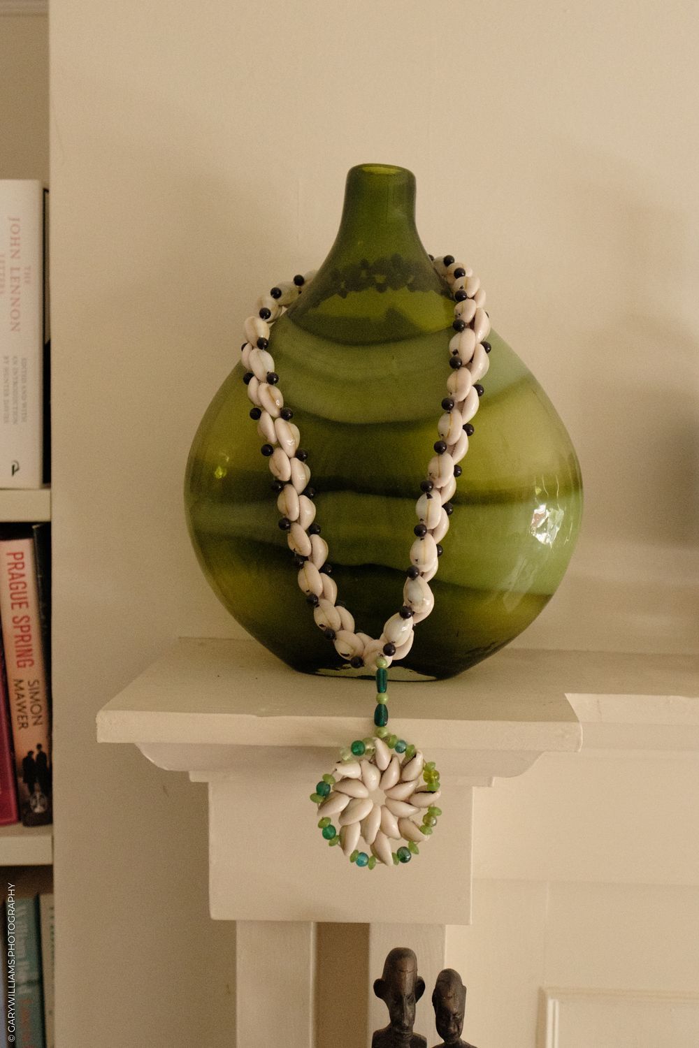 A green glass vase with a white beaded necklace draped around it and a circular pendant on display on a white shelf.