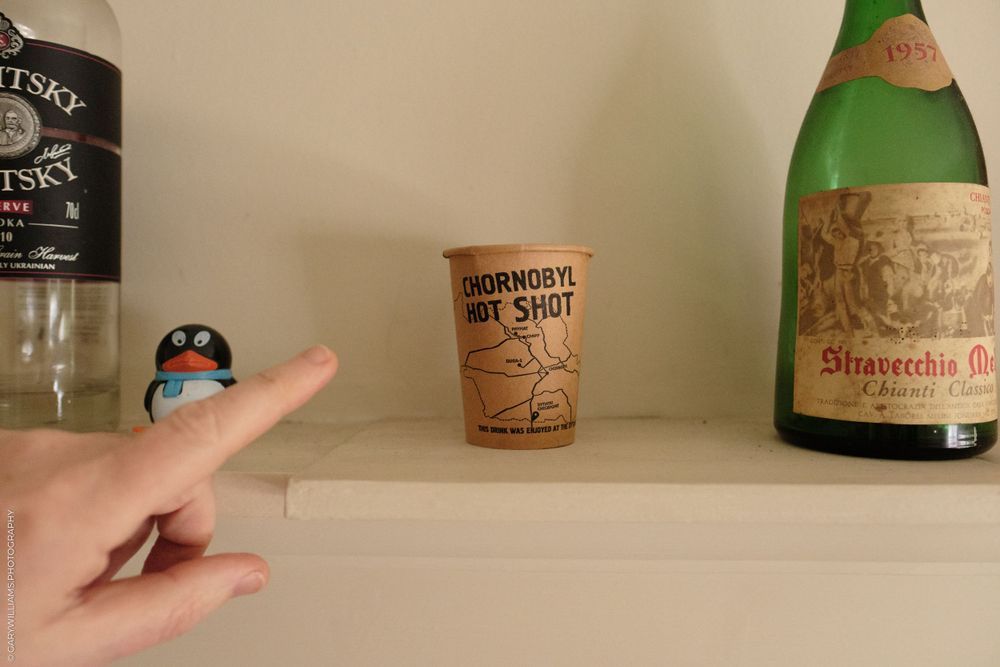 A finger points to a disposable coffee cup on a shelf between a dark bottle and a green glass bottle.