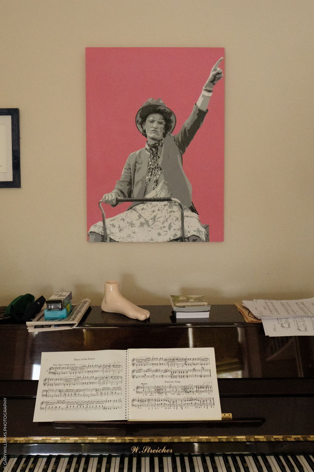 Sheet music sits on a piano under a pink and gray photo print, with a decorative shoe and books on the piano's wooden surface.