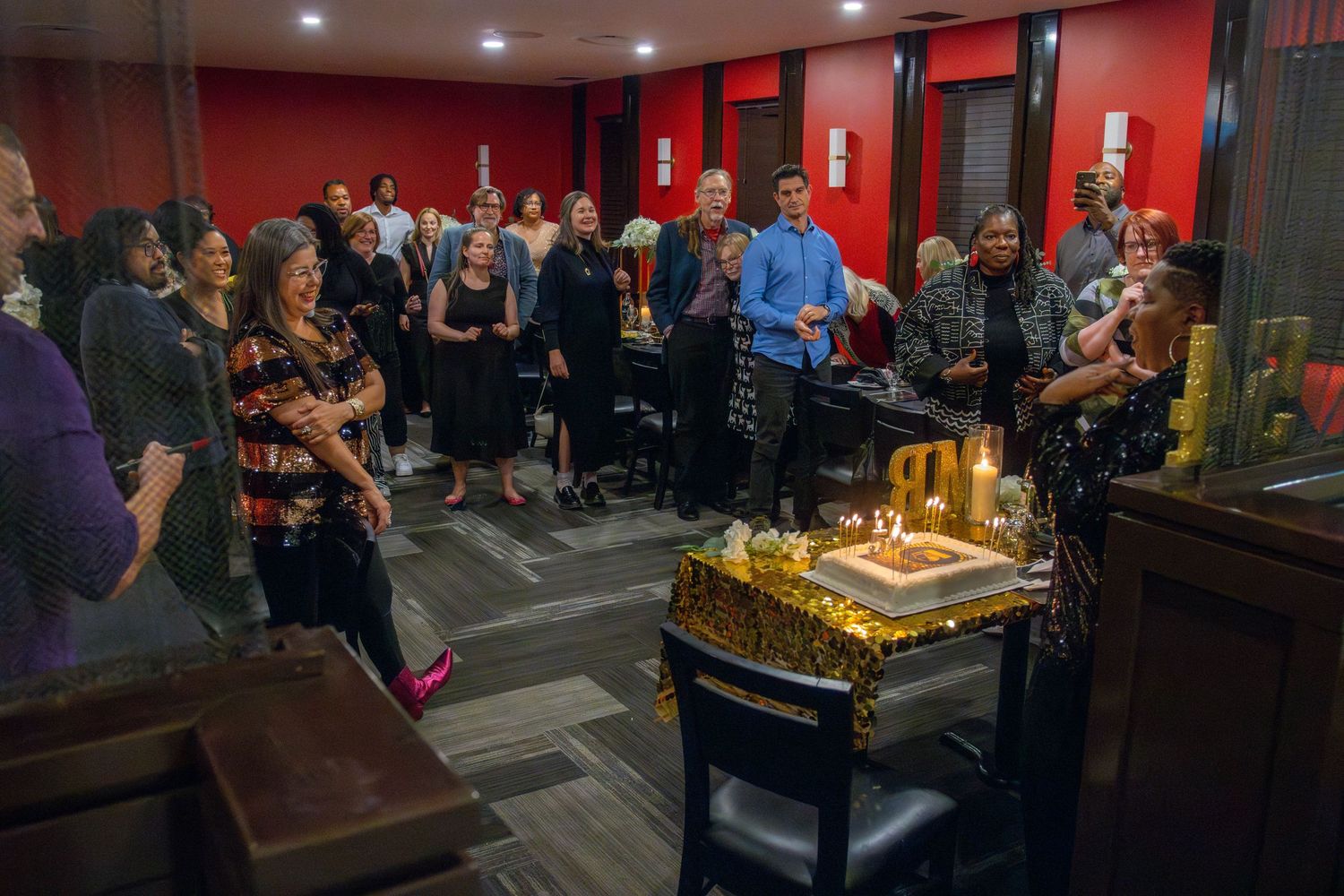 A crowd gathers around a cake with lit candles at an indoor celebration event with red walls.