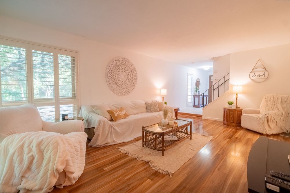Bright and airy living room with hardwood floors, plush couches, white decor, and natural lighting from large windows.