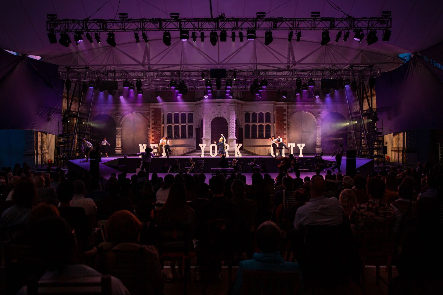 Wide shot of theater stage with purple lighting and audience visible in the foreground.
