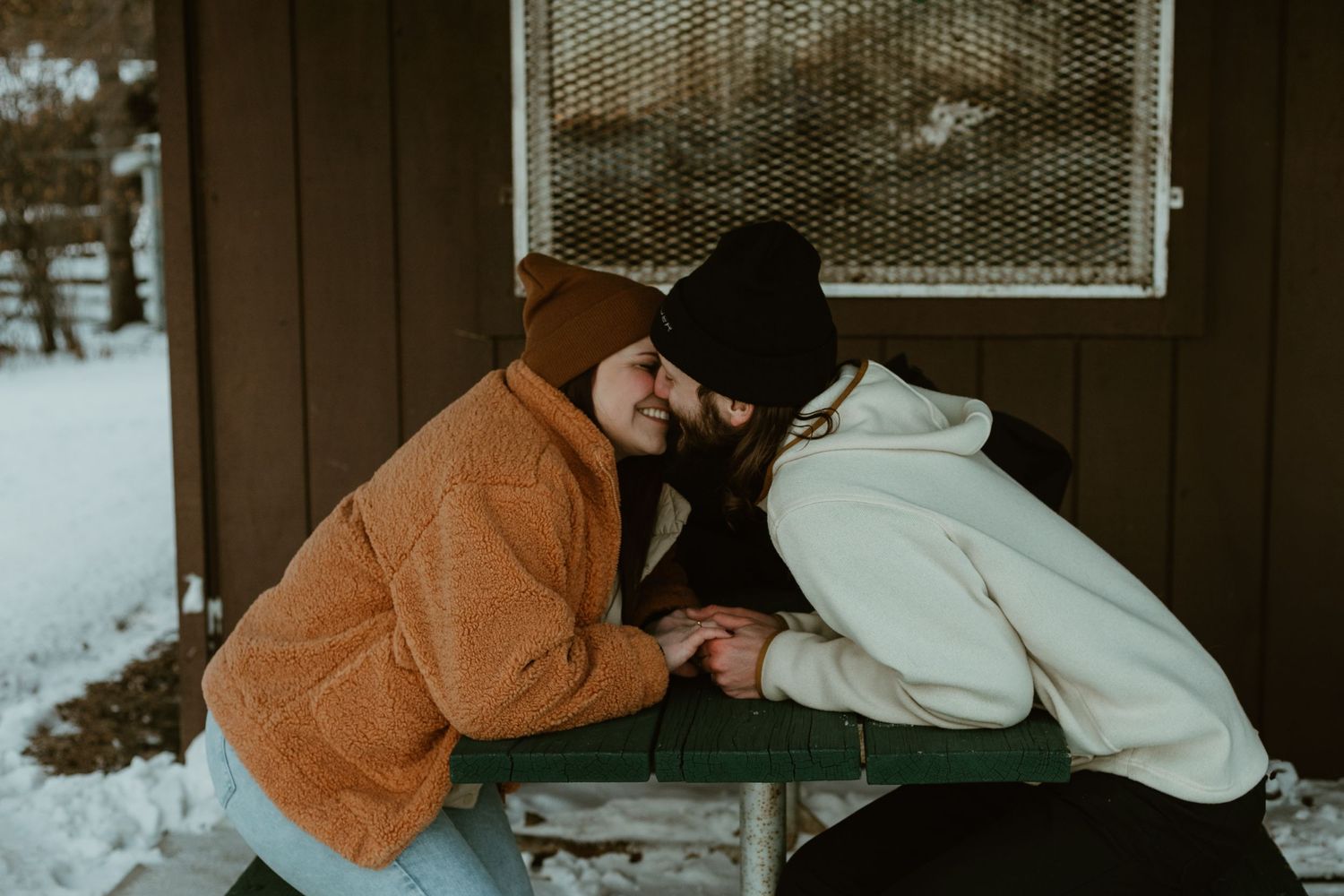 Two people in winter coats share an intimate moment on a wooden bench during a snowy day.