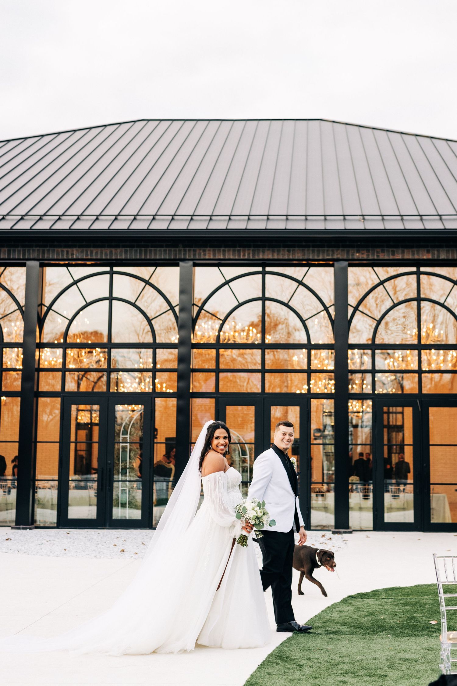 Wedding couple poses in front of elegant glass conservatory with arched windows and metal roof at sunset.