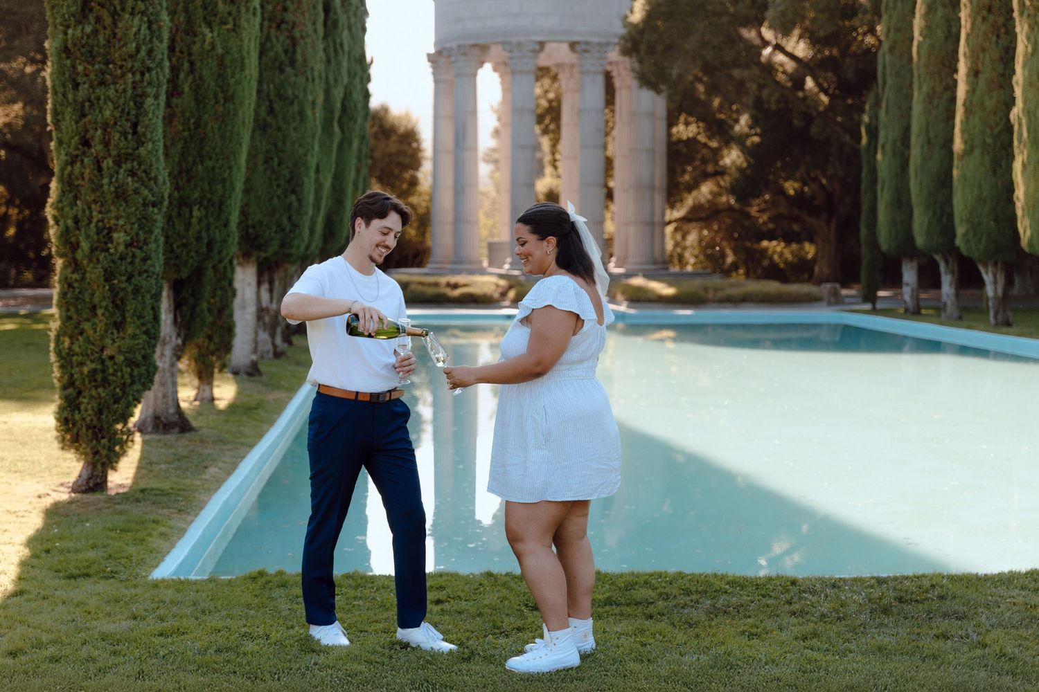 Couple dancing by reflective pool with classical columns and cypress trees in background.
