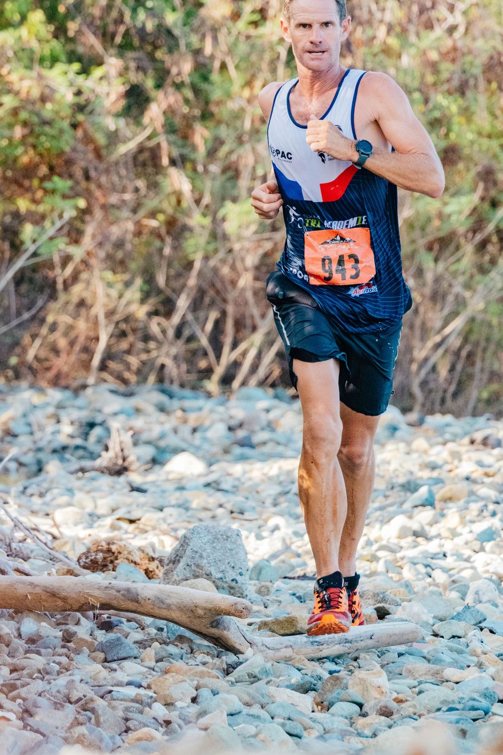 Trail runner navigating rocky terrain wearing blue tank top and shorts with race number in outdoor competition.