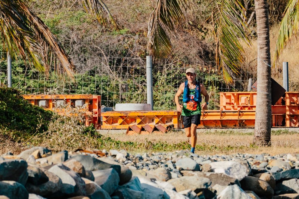 A runner jogs along a coastal path with wooden benches and palm trees beside a rocky shoreline.