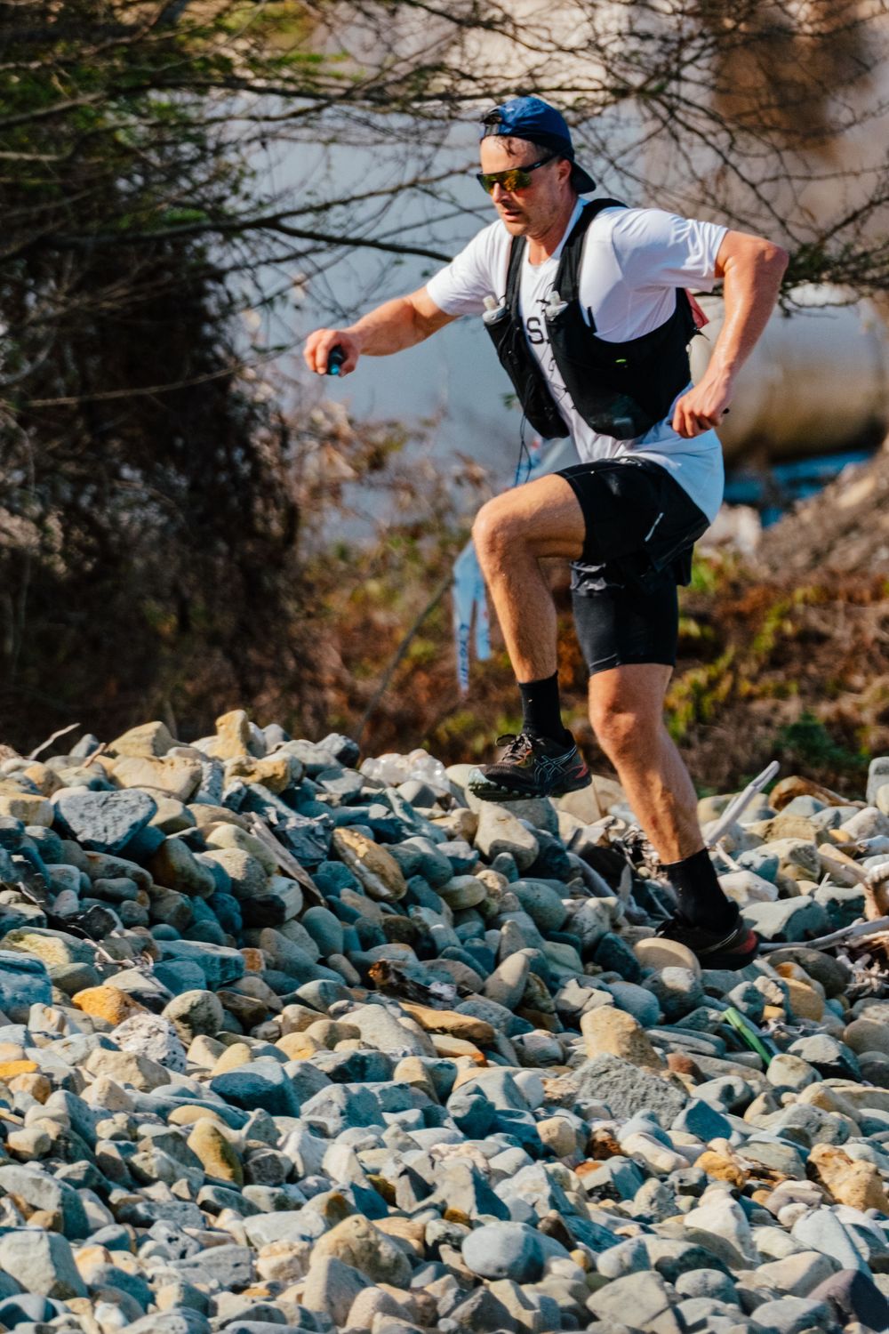 Trail runner traverses rocky terrain wearing sunglasses, blue cap, white shirt and black shorts in outdoor adventure setting.