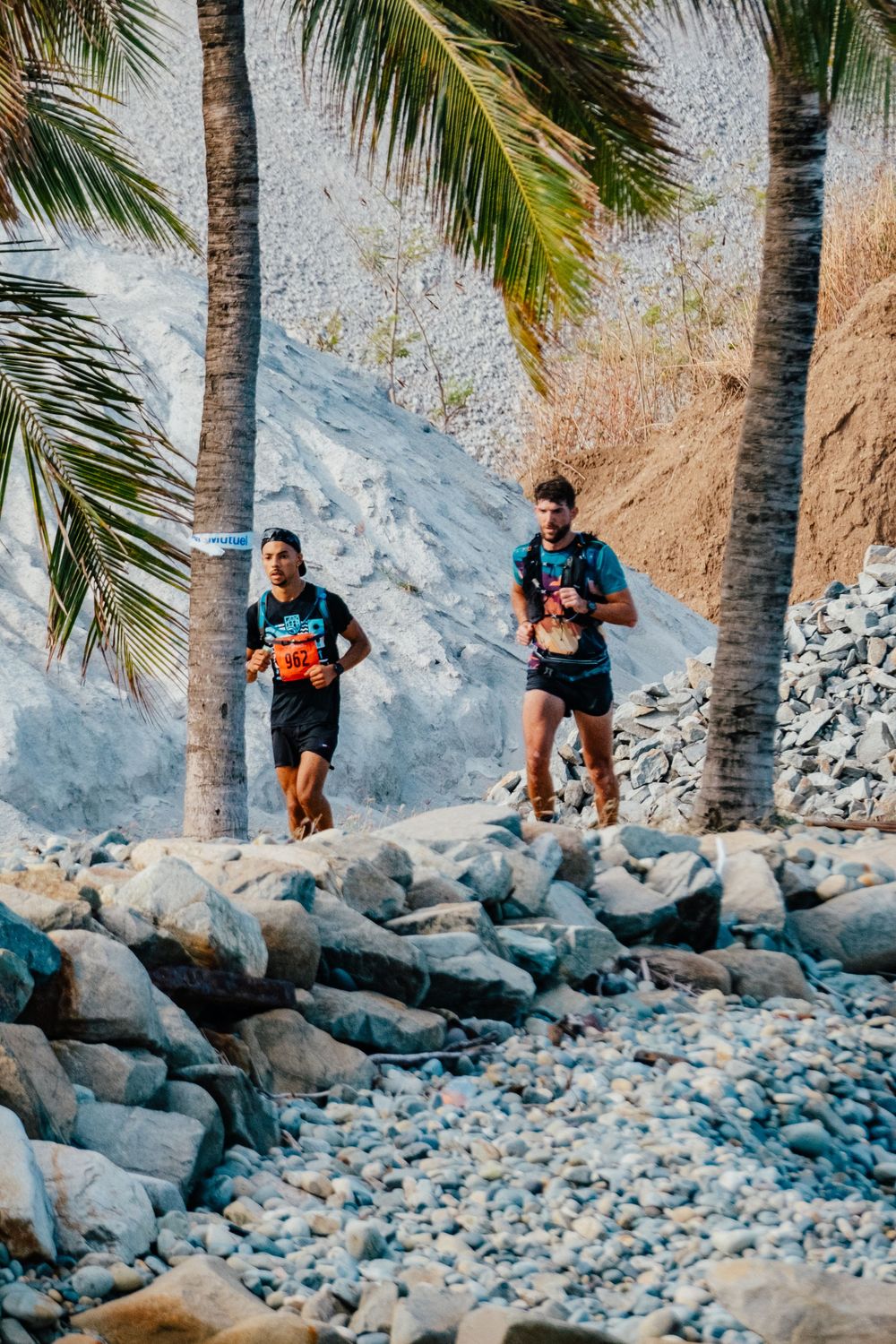Two trail runners navigate rocky terrain along a coastal path with palm trees and cliffs in the background.